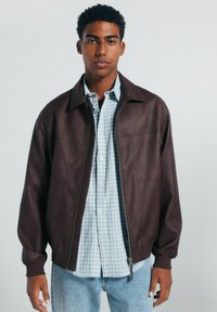 Braune Lederbomberjacke mit gerippten Bündchen und Reißverschluss, getragen über einem blau-weißen Karoshirt, kombiniert mit hellen Denim-Jeans.