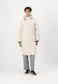 Polstret lang jakke i off-white, med hette, frontlommer og kviltet tekstur. Sammen med lyseblå jeans og svarte sneakers.