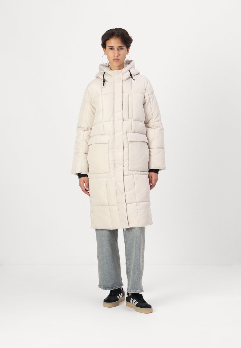 Polstret lang jakke i off-white, med hette, frontlommer og kviltet tekstur. Sammen med lyseblå jeans og svarte sneakers.