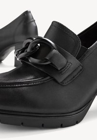 Mocassins noirs en cuir à talons avec un détail de chaîne épaisse à l'avant et des coutures décoratives, présentés sur fond blanc.