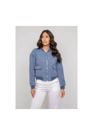 Femme aux longs cheveux bruns, vêtue d'une veste bomber bleue zippée et d'un pantalon blanc, se tenant devant un fond uni gris clair.