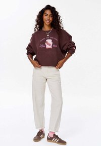 Hnědý oversized svetr s grafickým potiskem, bílými nápisy, krémové kalhoty a hnědé tenisky s bílými pruhy. Text obsahuje "Kde pes spí."