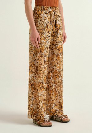 Woman wearing wide-leg pants with orange butterfly wing print, brown top, and beaded sandals with wooden oval detail.