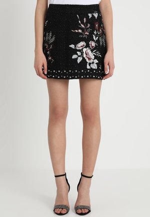 Mini-jupe noire avec broderie florale blanche et bordeaux et clous argentés le long de l'ourlet, portée avec des talons noirs à bride cheville.