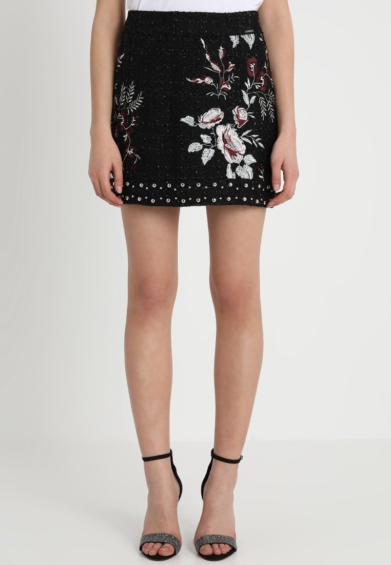 Mini-jupe noire avec broderie florale blanche et bordeaux et clous argentés le long de l'ourlet, portée avec des talons noirs à bride cheville.
