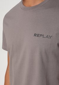 Gros plan d’une personne portant un t-shirt gris avec le mot "REPLAY" imprimé en noir sur la partie gauche de la poitrine.