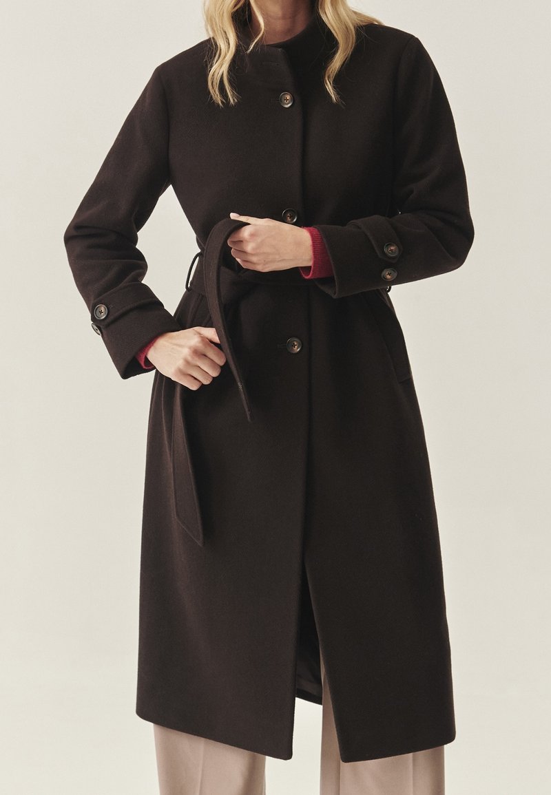 Femme attachant une ceinture sur un long manteau marron foncé à boutons, portant un pantalon beige et des manches de pull rouge visibles aux poignets.