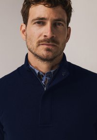 Marineblauwe gebreide trui met een opstaande kraag en ritssluiting, gedragen over een blauw overhemd met kraag, voorzien van knopen en een textuurstof.