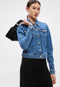 Modrá rifľová bunda s krátkym strihom, zapínaná na gombíky a s dvoma náprsnými vreckami. Čierna kabelka s textúrovaným povrchom držaná v ruke modelky.