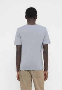 Άνδρας που φοράει απλό ανοιχτό γκρι κοντομάνικο μπλουζάκι και μπεζ παντελόνι, στέκεται μπροστά σε ένα λευκό τοίχο με χαλαρά τα χέρια στα πλάγια.