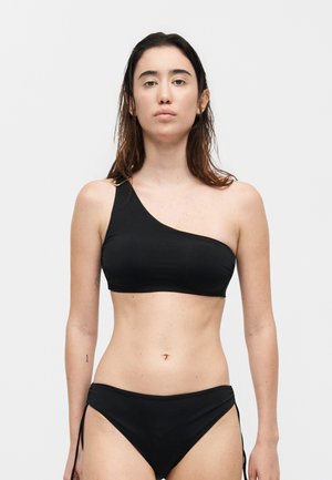 Γυναίκα με μακριά σκούρα μαλλιά που φοράει μαύρο μπικίνι τοπ με έναν ώμο και ταιριαστό κάτω μέρος μαγιό σε απλό ανοιχτό φόντο.