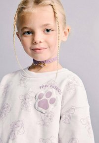 Sweat-shirt blanc avec des motifs de chiens dessinés en violet clair, un patch en relief en forme d'empreinte de patte, et le texte « PAW PATROL ». Collier ras-du-cou violet avec un charme.