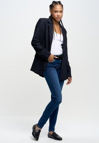 Crni sako s trakama, bijela majica bez rukava, plave uske traperice i crne loafersice. Model ima pletenu kosu i pozira s jednom rukom na kuku.