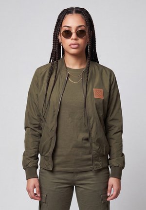 Woman with long braids wearing round sunglasses, layered gold necklaces, an olive green jacket with a patch, matching shirt, and cargo pants.