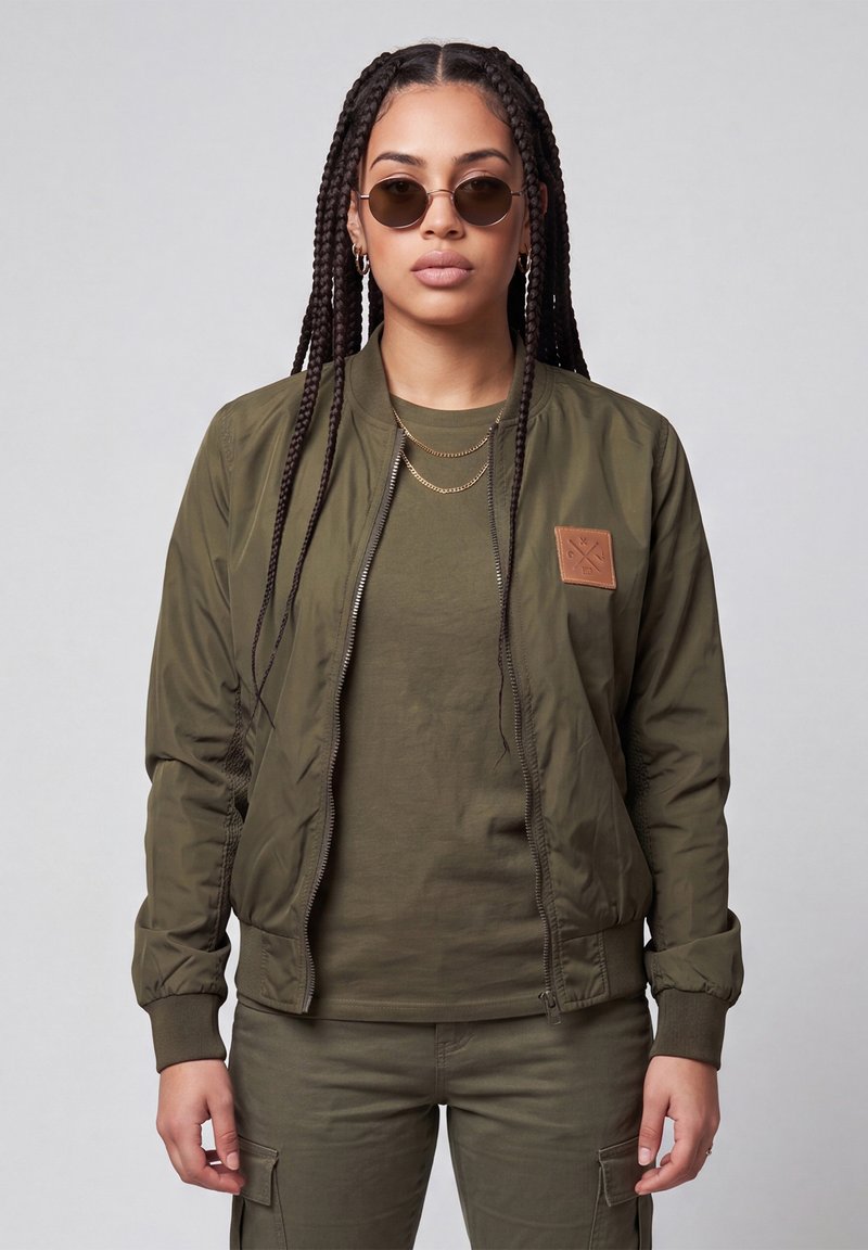 Woman with long braids wearing round sunglasses, layered gold necklaces, an olive green jacket with a patch, matching shirt, and cargo pants.