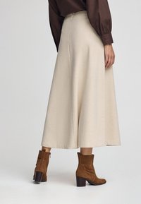 Kvinde iført en cremefarvet midi-skjorte, brun langærmet bluse og brune ruskind ankelstøvler med blokhæv, stående på en ensfarvet baggrund.