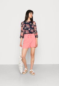 Blusa floral com mangas transparentes em azul-marinho, combinada com calções cor-de-rosa. Acessórios incluem uma mala de mão branca e sandálias com tira.