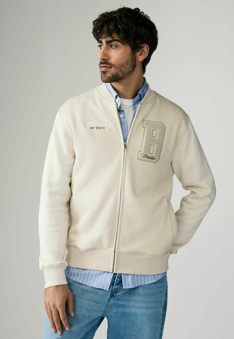 Beige zip-up sweatshirt med ribbet krave, der har en stor "B" applikation og broderet tekst. Bæres over en lyseblå stribet skjorte.
