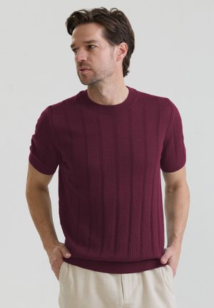 Mand iført en bordeaux tekstureret kortærmet sweater og lyse beige bukser, der kigger til siden med hænderne i lommerne mod en ensfarvet baggrund.