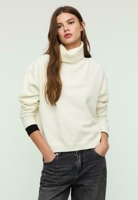 Cremfarbener Rollkragensweater aus weichem Stoff mit langen Ärmeln, schwarzen Bündchenakzenten und lockerer Passform, kombiniert mit dunkelblauen Jeans.