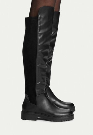 Black knee-high boots with smooth leather front and suede back panels worn with black hosiery on a light background.