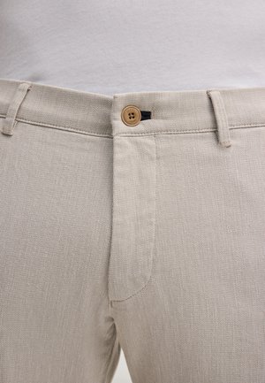 Primo piano della vita di pantaloni beige testurizzati con un bottone marrone e passanti per cintura, indossati con una camicia bianca infilata dentro.