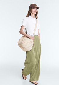 Femme portant une casquette marron, un polo blanc, un pantalon large vert olive, des tongs, portant un sac d'épaule matelassé beige, marchant sur fond blanc.