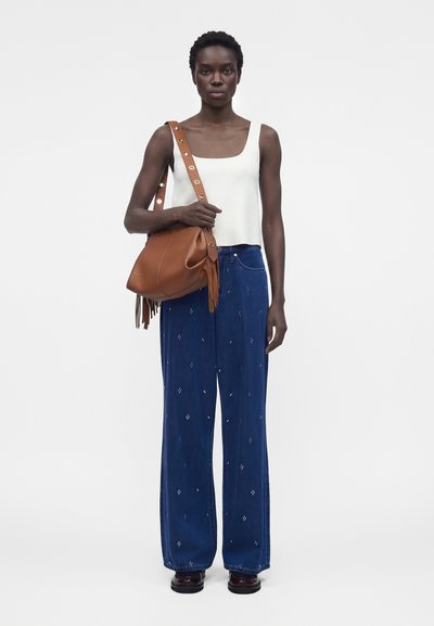 Γυναίκα που φοράει λευκό αμάνικο τοπ και φαρδιά μπλε παντελόνια, κρατώντας καφέ δερμάτινη τσάντα ώμου με διακοσμητικά κρόσσια σε λευκό φόντο.