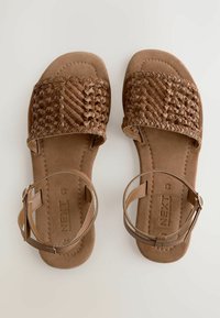 Hnědé tkané sandály s plochou podrážkou, nastavitelnými popruhy kolem kotníků a měkkým semišovým povrchem. Obsahují zapletený design na vršku a minimální kovové doplňky.