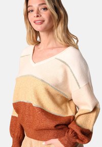 Pull à col en V multicolore dans des teintes de crème, beige et orange rouille. Texture douce avec des coutures contrastées et des manches bouffantes.