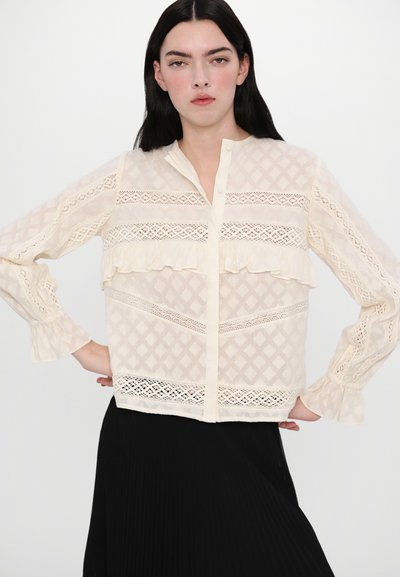 Blusa de crema claro con intrincados encajes y patrones de diamantes, con botones al frente, acentos con volantes en las mangas y el cuerpo, textura semitranslúcida.