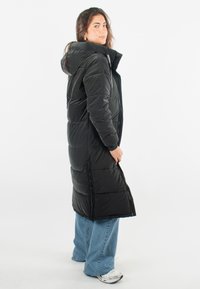 Manteau matelassé noir avec capuche, longueur longue, texture matelassée et zip sur toute la longueur ; associé à un jean en flares bleu et des chaussures de sport.
