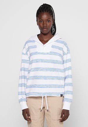 Junge Frau mit geflochtenem Haar, die einen weißen Hoodie mit dünnen blauen Streifen und beigefarbene Cargo-Hosen trägt, stehend vor einem schlichten Hintergrund.