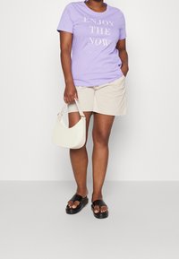 Lilla grafisk t-shirt med teksten "Nyd nuet", beige shorts, sorte slidesandaler og en hvid håndtaske. Enkel design, afslappet pasform.