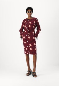 Burgundy blommig klänning med långa ärmar, knytmidja och mönster av rosa och vita blommor. Kombinerad med tjocka svarta sandaler.