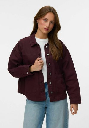 Kvinde med langt brunt hår iført en mørkelilla knappet jakke over en hvid skjorte og lyseblå jeans mod en ensfarvet baggrund.