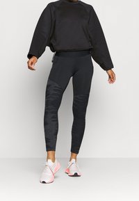 Sweat-shirt noir avec des manches texturées, associé à des leggings noirs présentant un motif subtil. Des chaussures de sport blanches et roses complètent la tenue.