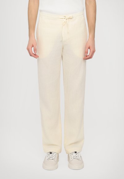 Pantaloni in lino beige chiaro con una vestibilità rilassata, dotati di una vita con cordoncino, gamba dritta e una texture sottile. Abbinati a sneakers bianche.