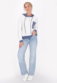Felpa bianca con cappuccio e dettagli a strisce blu, tre bottoni e tasca frontale; abbinata a jeans blu chiaro e sneakers bianche.