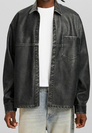 Mann trägt ausgewaschenes schwarzes Hemd mit Knöpfen über weißem T-Shirt und hellblauen Jeans, steht vor schlichtem Hintergrund.