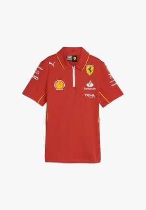 Czerwona koszulka polo z żółtymi akcentami; posiada zamek błyskawiczny przy kołnierzy oraz wiele logo, w tym Ferrari, Shell i Puma, na przodzie i rękawach.