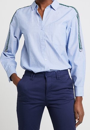 Femme portant une chemise bleu clair à boutons avec des détails rayés verts sur les manches et un pantalon taille haute bleu marine, une main dans la poche.