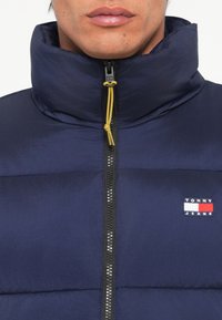 Marineblauer Puffer-Jacke mit hohem Kragen, schwarzem Reißverschluss und gelbem Zug; mit kleinem Tommy Jeans Logo auf der Brust. Strukturiertes Material.