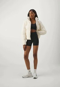 Λευκό, φουντωτό μπουφάν με ψηλό γιακά φορεμένο πάνω από μαύρο σπορ σουτιέν και ταιριαστά σορτς; μοντέλο με λευκά παπούτσια, στέκεται μπροστά σε ανοιχτό φόντο.