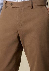 Primo piano di pantaloni marroni fissati con un solo bottone, indossati con una camicia a righe infilata dentro, che mostrano le tasche frontali e i passanti per la cintura.