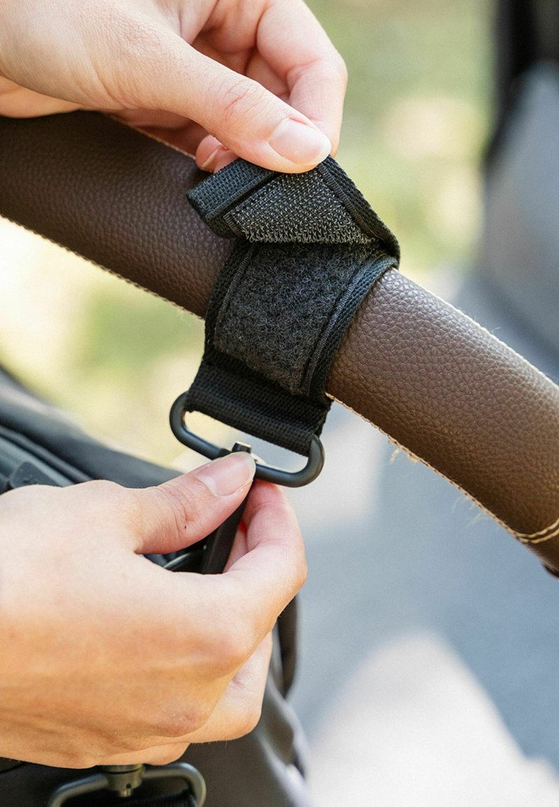 Justerbar sort velcro-rem, der sikrer et brunt tekstureret håndtag med en metalring til fastgørelse, holdt af en hånd.