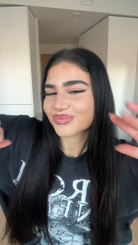 Mladá žena s dlhými čiernymi vlasmi, ktorá má na sebe tmavé tričko s potlačou, usmieva sa a dvíha ruky v interiéri, v pozadí biele skrinky.