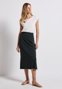 Bílé tričko s krátkým rukávem, které je kombinováno s dlouhou, tmavě zelenou, přiléhavou sukní s asymetrickým střihem a hladkým povrchem. Oblečení doplňují sandály.