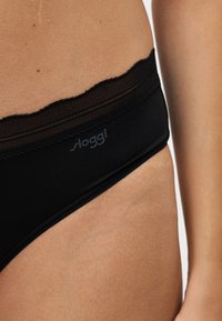 Крупен план на човек, носещ черно бельо с дантелена ивица и лого "sloggi" на ластика в областта на ханша.