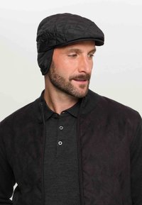 Cappello nero trapuntato con orecchiette, dalla parte superiore arrotondata e dal design elegante. Indossato con una giacca scura e una polo grigia. Texture liscia.